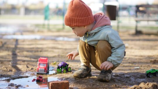 Kleiner Junge hockt in einer Schlammpfütze und spielt mit seinen Plastikspielzeugautos.