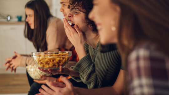 Freunde entspannen sich gemeinsam zu Hause, schauen fern und essen Snacks. Sie sehen sich den Eurovision Song Contest an.