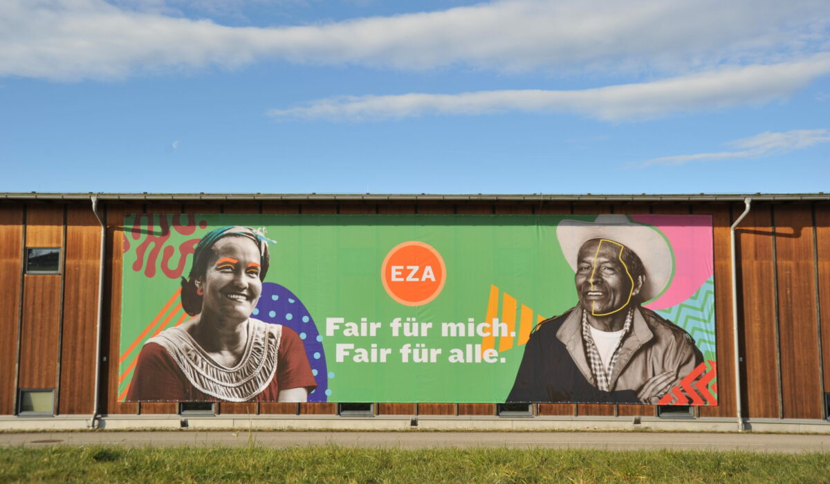 Werbebanner von EZA fairer Handel auf Gebäude mit Bildern zweier Menschen links und rechts