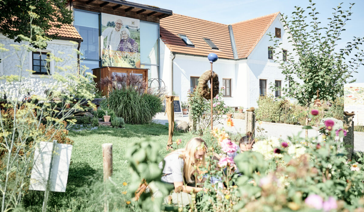 Sonnentor Firmengebäude hinter blühenden Garten, zwei Frauen bücken sich hinunter zu Pflanzen