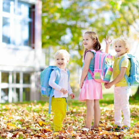 Drei Kinder mit Rucksäcken und Schultasche vor einem Gebäude unter einem Baum mit Falllaub