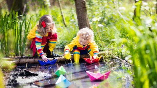 Zwei Kinder spielen mit Papierbooten am Bach.