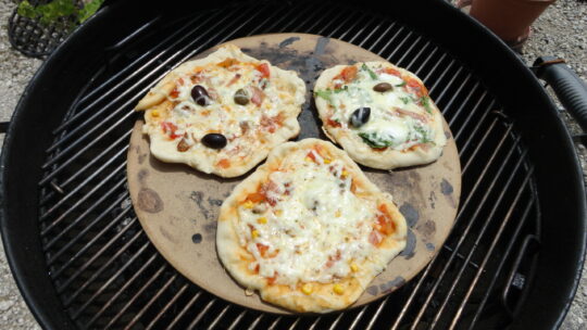 Drei kleine Pizzen auf einem Pizzastein auf einem Griller.
