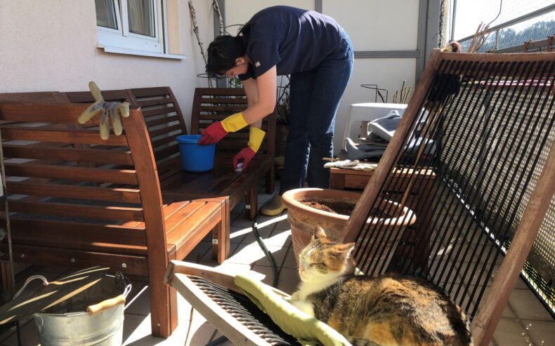 Frau putz Balkonmöbeln mit Kübel und Schwamm und eine Katze liegt daneben in einem Liegestühl