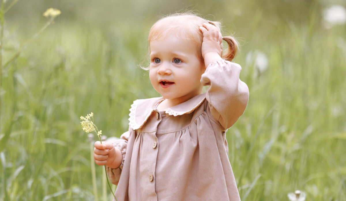 Ein Kind in einem hellbraunen Kleid steht auf einem Feld und hält eine kleine Blume in der Hand.