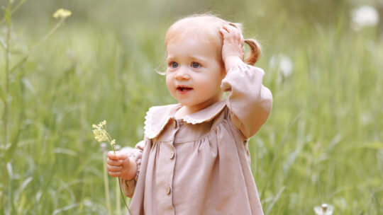Ein Kind in einem hellbraunen Kleid steht auf einem Feld und hält eine kleine Blume in der Hand.