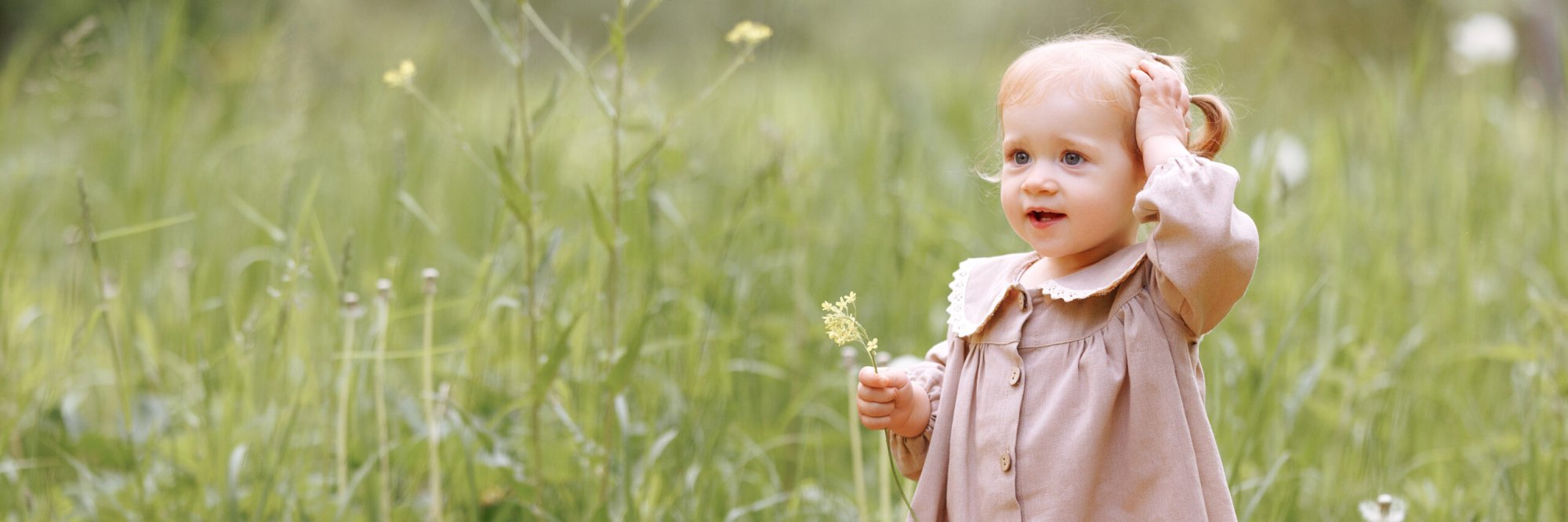 Ein Kind in einem hellbraunen Kleid steht auf einem Feld und hält eine kleine Blume in der Hand.