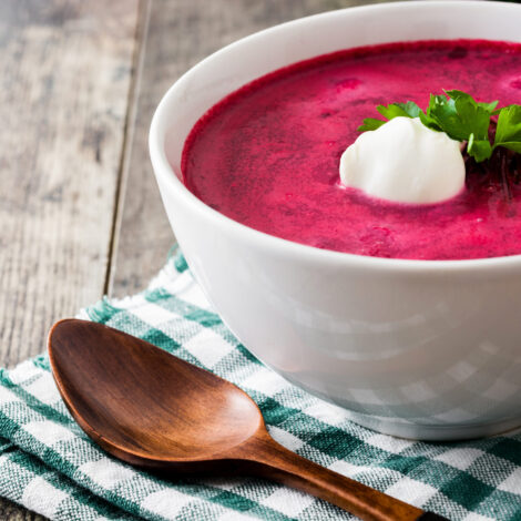 Rote Rübensuppe in einer weißen Schale mit Rahm und Petersilientopping auf einem grün, weiß karierten Tuch und einem Holzlöffel