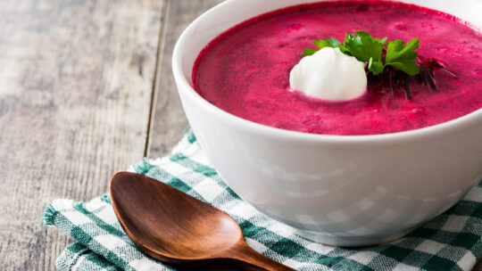 Rote Rübensuppe in einer weißen Schale mit Rahm und Petersilientopping auf einem grün, weiß karierten Tuch und einem Holzlöffel