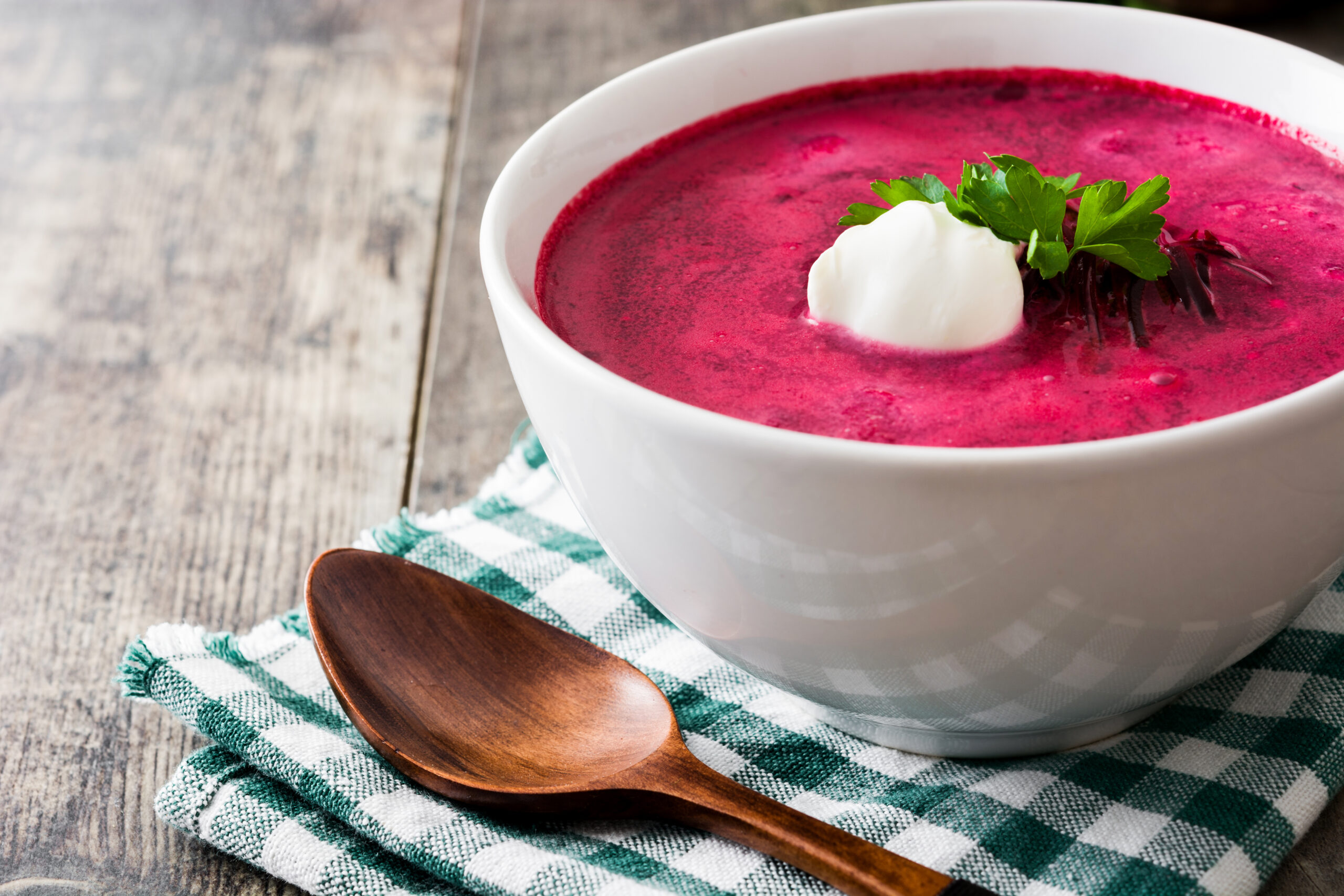 Rote Rübensuppe in einer weißen Schale mit Rahm und Petersilientopping auf einem grün, weiß karierten Tuch und einem Holzlöffel