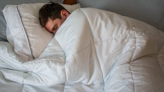 Junger Mann schläft im Bett unter einer kuscheligen Bettdecke.