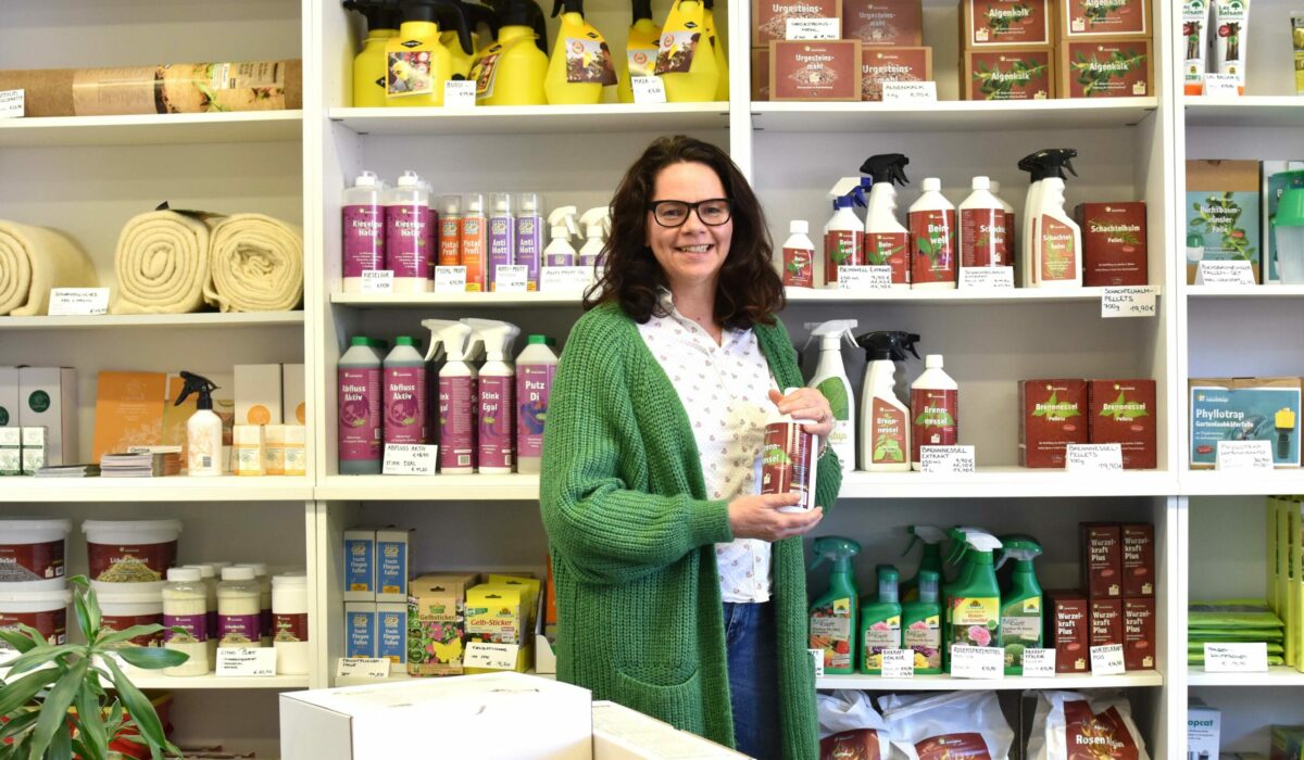 DI Sabine Pleininger, Geschäftsführerin von biohelp Garten & Bienen GmbH mit natürlichem Düngemittel in Flasche in der Hand, steht vor Regal mit einigen ihrer Produkte.