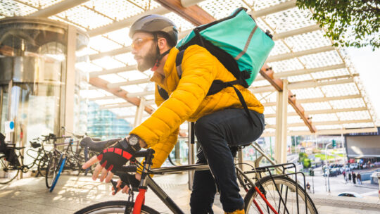 Fahrradkurier mit gelber Jacke und grünem Transportrucksack liefert Pakte mit dem Fahrrad an Kunden
