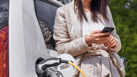 Frau schaut auf ihr Smartphone beim Laden eines E-Autos