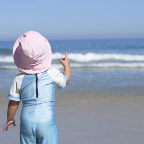 Kleinkind mit rosafarbenem Sonnenhut und hellbauen Sonnenschutzbody steht am Sandstrand, blickt auf den Horizont über dem Meer, Rückansicht