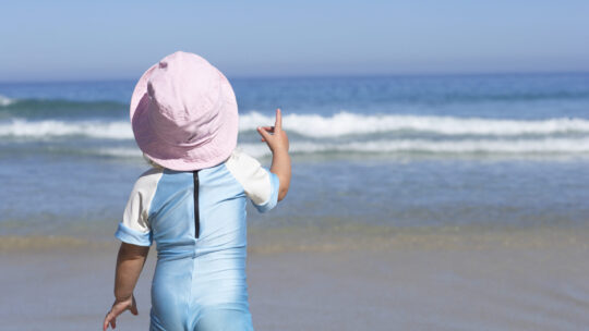 Kleinkind mit rosafarbenem Sonnenhut und hellbauen Sonnenschutzbody steht am Sandstrand, blickt auf den Horizont über dem Meer, Rückansicht