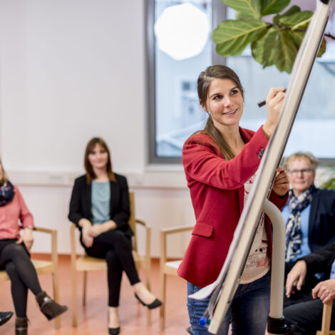 Eine Vortragende schreibt auf ein Flipchart und die Kursteilnehmer und Kursteilnehmerinnen sitzen im Halbkreis