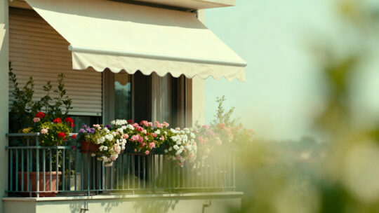 Foto, heißer Sommertag, Balkon mit Blumen und Markise und heruntergelassenen Außenrollos zur Abkühlung