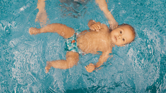 Baby mit Schwimmwindel wird von Mutter im Pool über Wasser gehalten