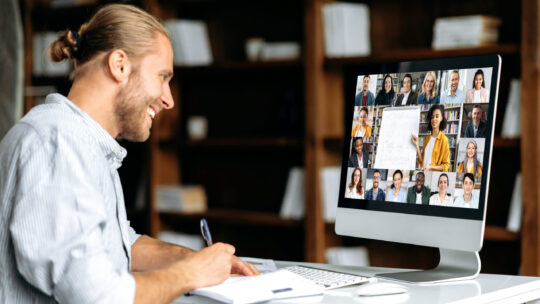 Junger Mann sitzt vor einem PC nimmt an einem online-Seminar teil und schreibt sich Informationen in ein Notizbuch