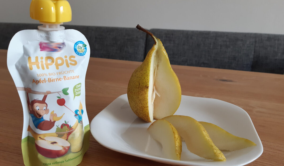 Aufgeschnittene Birne auf einem Teller neben einer gekauften Packung Fruchtmus im Plastikbeutel.