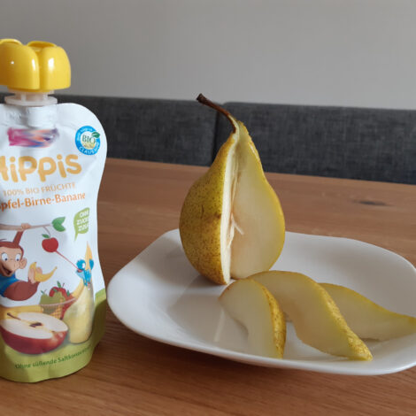 Aufgeschnittene Birne auf einem Teller neben einer gekauften Packung Fruchtmus im Plastikbeutel.