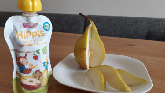 Aufgeschnittene Birne auf einem Teller neben einer gekauften Packung Fruchtmus im Plastikbeutel.