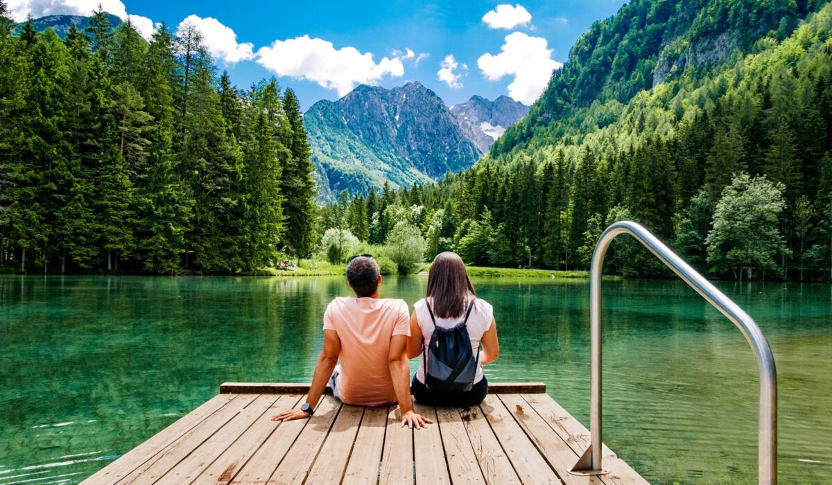 Junges Paar sitzt auf einem Holzsteg an einem grünen See, umgeben von Wäldern und Bergen mit blauem Himmel und weißen Wolken darüber.