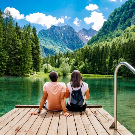 Junges Paar sitzt auf einem Holzsteg an einem grünen See, umgeben von Wäldern und Bergen mit blauem Himmel und weißen Wolken darüber.