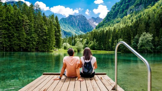 Junges Paar sitzt auf einem Holzsteg an einem grünen See, umgeben von Wäldern und Bergen mit blauem Himmel und weißen Wolken darüber.