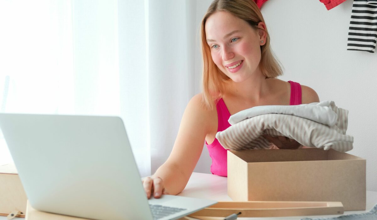 Frau sitzt vor ihrem Laptop und verpackt Kleidung in eine Schachtel.