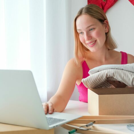 Frau sitzt vor ihrem Laptop und verpackt Kleidung in eine Schachtel.