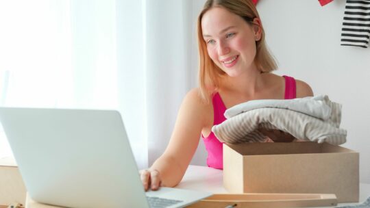 Frau sitzt vor ihrem Laptop und verpackt Kleidung in eine Schachtel.