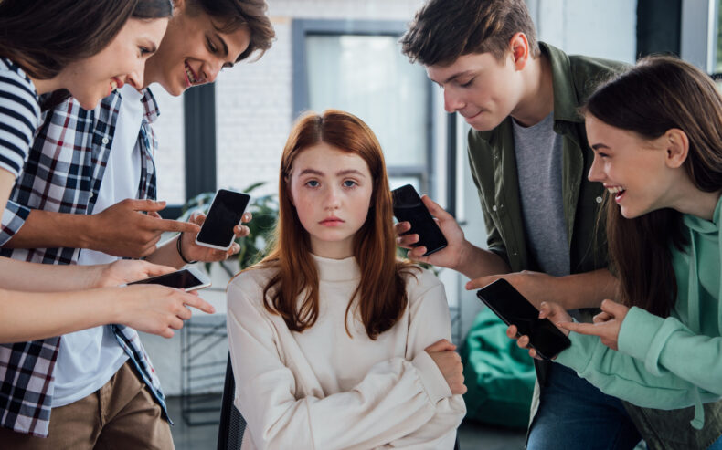 Ein Mädchen verzichtet auf ihr Smartphone und ihre Freunde stehen im Kreis um sie und zeigen lachend auf ihre smartphones