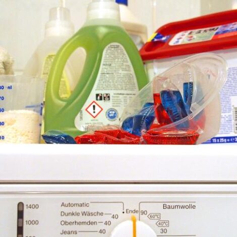 Auf einer Waschmaschine stehen neben bunten Handtüchern ein Beecher mit Pulverwaschmittel, eine Flasche Flüssigwaschmittel, rote und blaue Waschbeutel und ein Dose mit Spezialwaschmittel