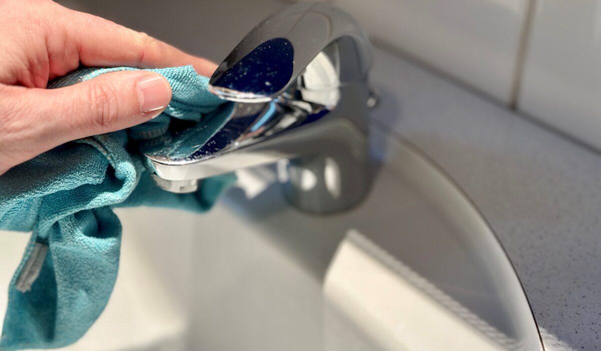 Eine Hand mit einem blauen Microfasertuch putzt einen Wasserhahn im Badezimmer
