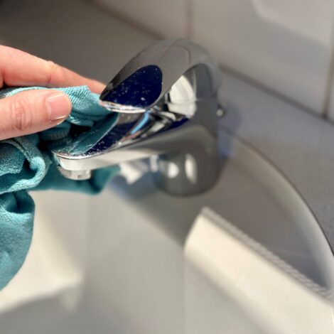 Eine Hand mit einem blauen Microfasertuch putzt einen Wasserhahn im Badezimmer