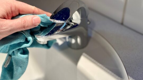 Eine Hand mit einem blauen Microfasertuch putzt einen Wasserhahn im Badezimmer