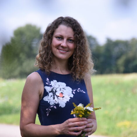 Elisabeth Teufner vor grüner Wiese und Laubbäumen mit Naturpflanzen in den Händen
