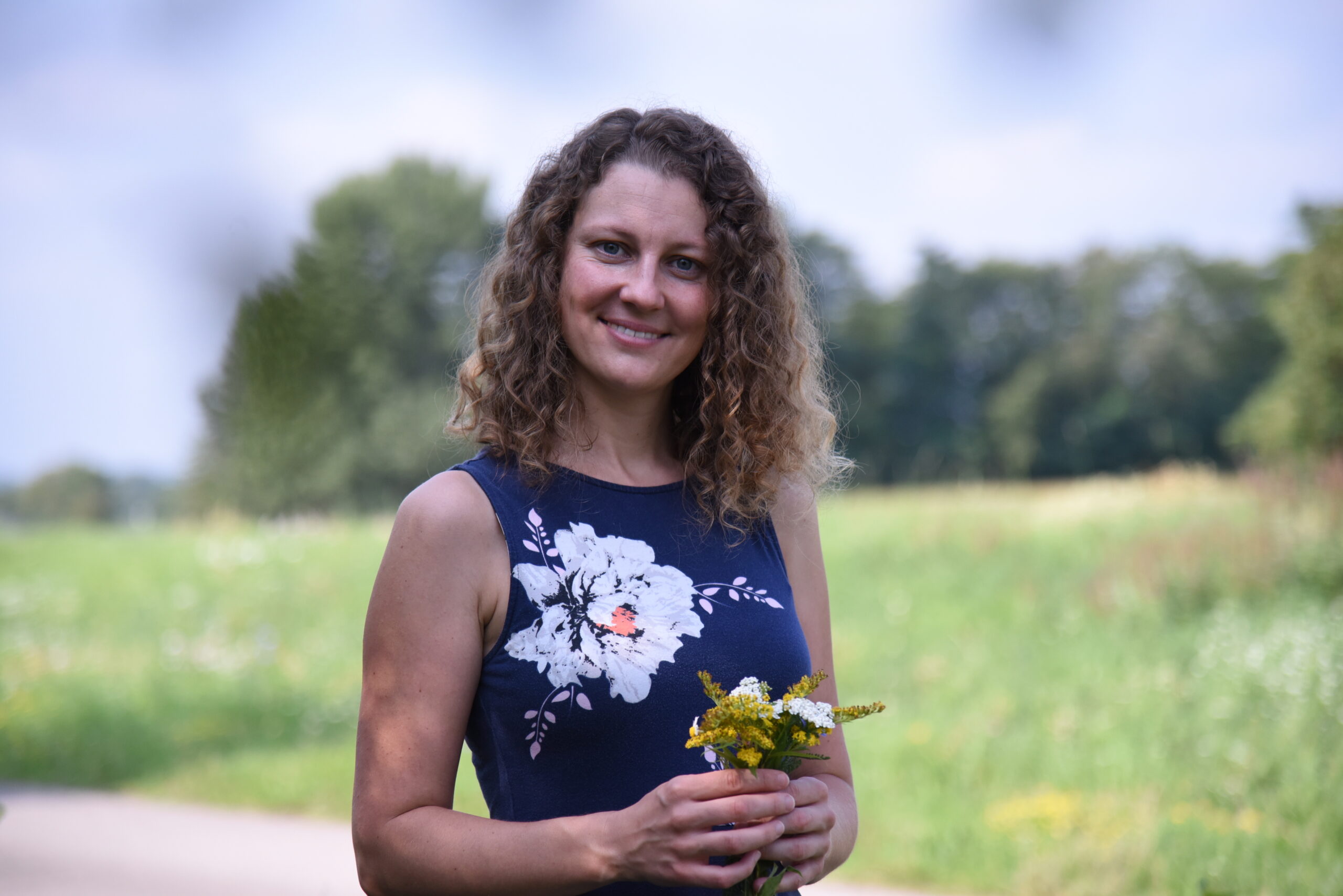 Elisabeth Teufner vor grüner Wiese und Laubbäumen mit Naturpflanzen in den Händen