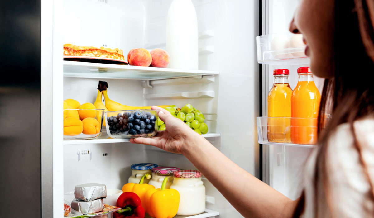 Frau nimmt sich eine Kiste Blaubeere aus dem offenen Kühlschrank voller Obst, Gemüse, Bananen, Pfirsiche und Joghurt.