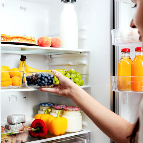 Frau nimmt sich eine Kiste Blaubeere aus dem offenen Kühlschrank voller Obst, Gemüse, Bananen, Pfirsiche und Joghurt.