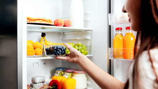 Frau nimmt sich eine Kiste Blaubeere aus dem offenen Kühlschrank voller Obst, Gemüse, Bananen, Pfirsiche und Joghurt.